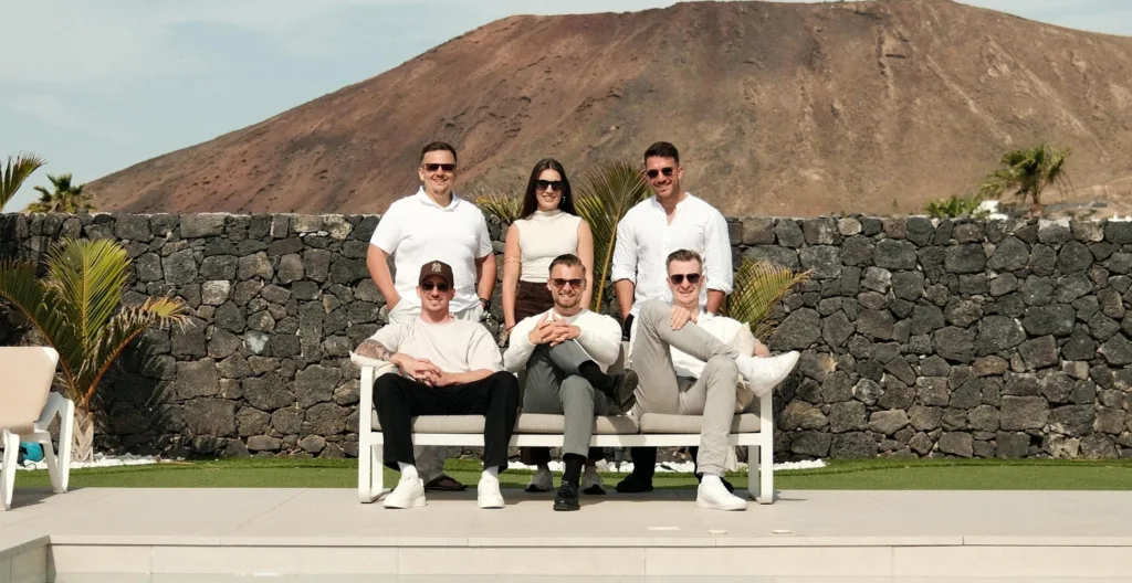 Das Team von BetonGold Immobilien posiert auf einer Terrasse am Pool vor einer Steinmauer und einer kargen Vulkanlandschaft.