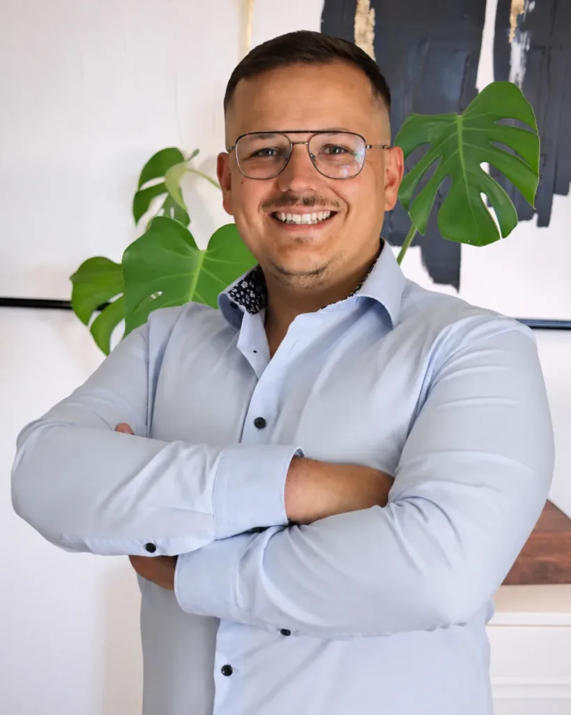 Alessandro lächelt im Büro von BetonGold Immobilien, mit verschränkten Armen vor einer modernen Wanddekoration und Pflanzen