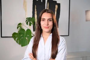 Sonja Gerst, Ansprechpartnerin bei BetonGold Immobilien, lächelt mit verschränkten Armen vor einer modernen Wanddekoration