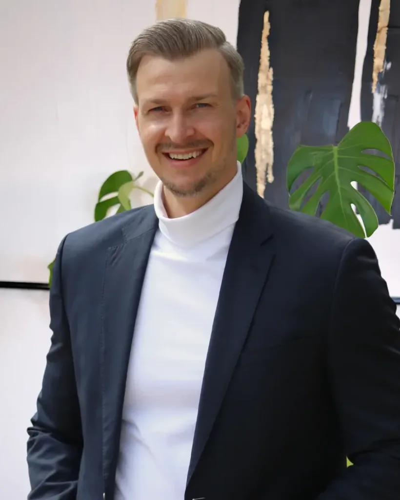 Tobias Heuse lächelt in einem schicken Outfit vor einem modernen Designer-Hintergrund mit Pflanzen, der professionalen Atmosphäre vermittelt
