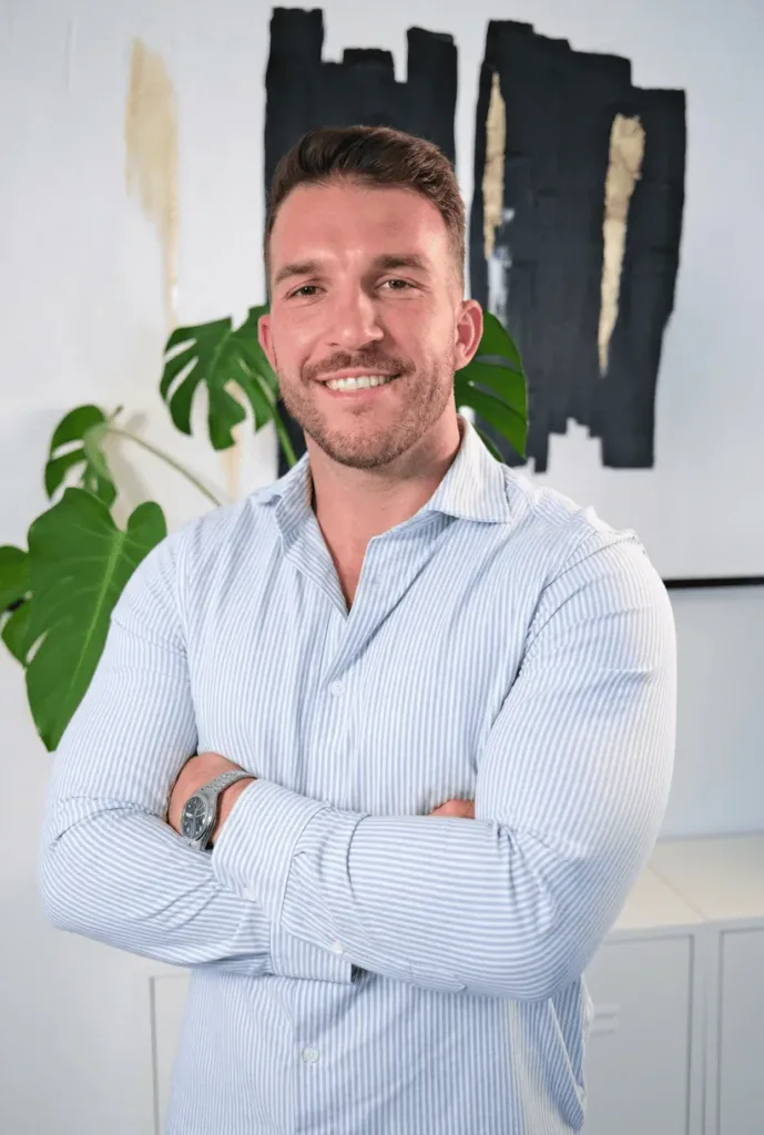 Johannes steht mit verschränkten Armen lächelnd vor einer modernen, minimalistischen Wandgestaltung, die eine positive und einladende Atmosphäre vermittelt